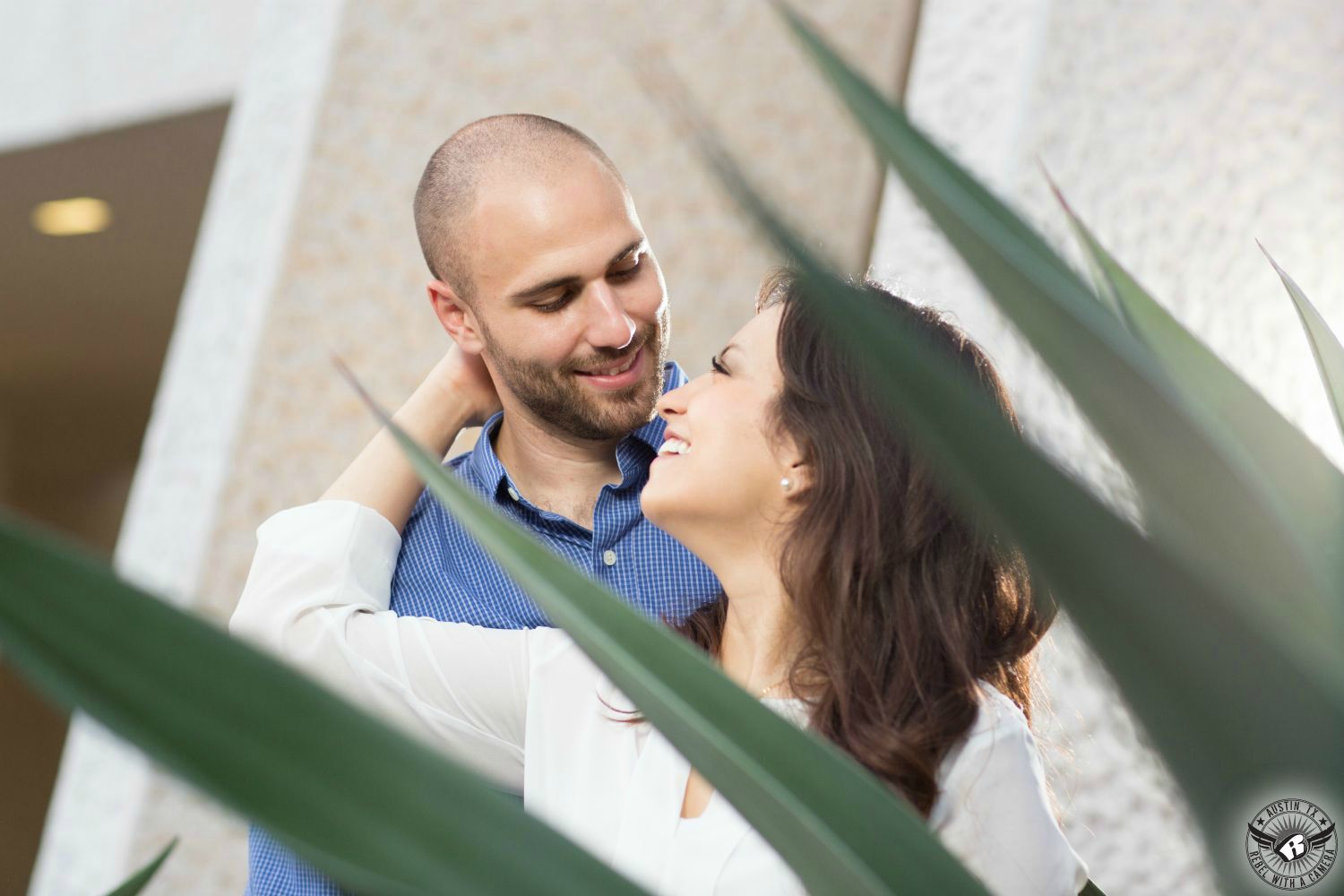 Wavy dark haired Latino girl in a white blouse looks up smiling at a guy with a short beard wearing a blue and white checked shirt with a succulent agave plant in the foreground obscuring the couple behind spiky leaves at the Mexican American Cultural Center in this elated engagement image in Austin, Texas.